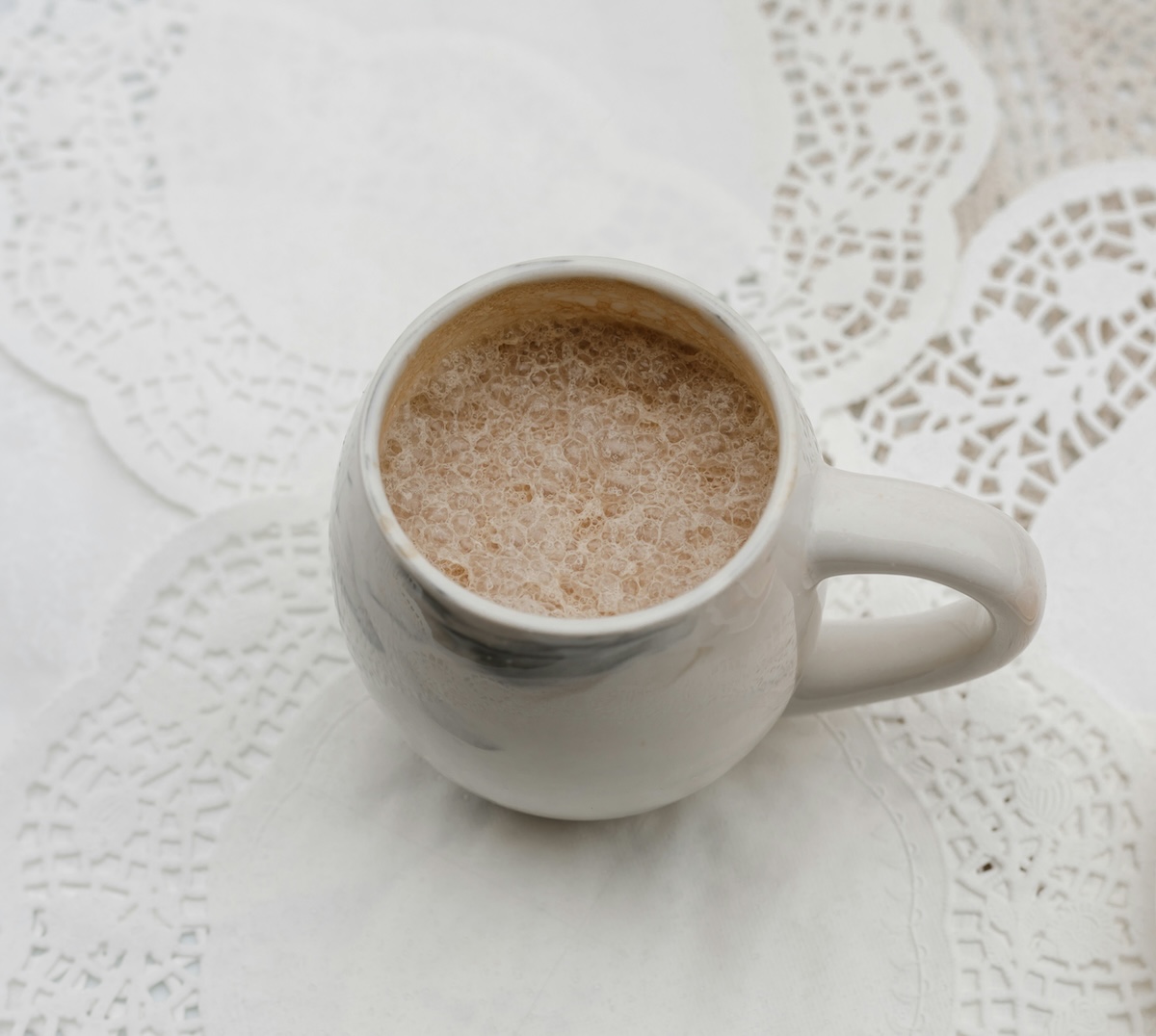 Overhead view of frothy hot chocolate in white mug on lace tablecloth